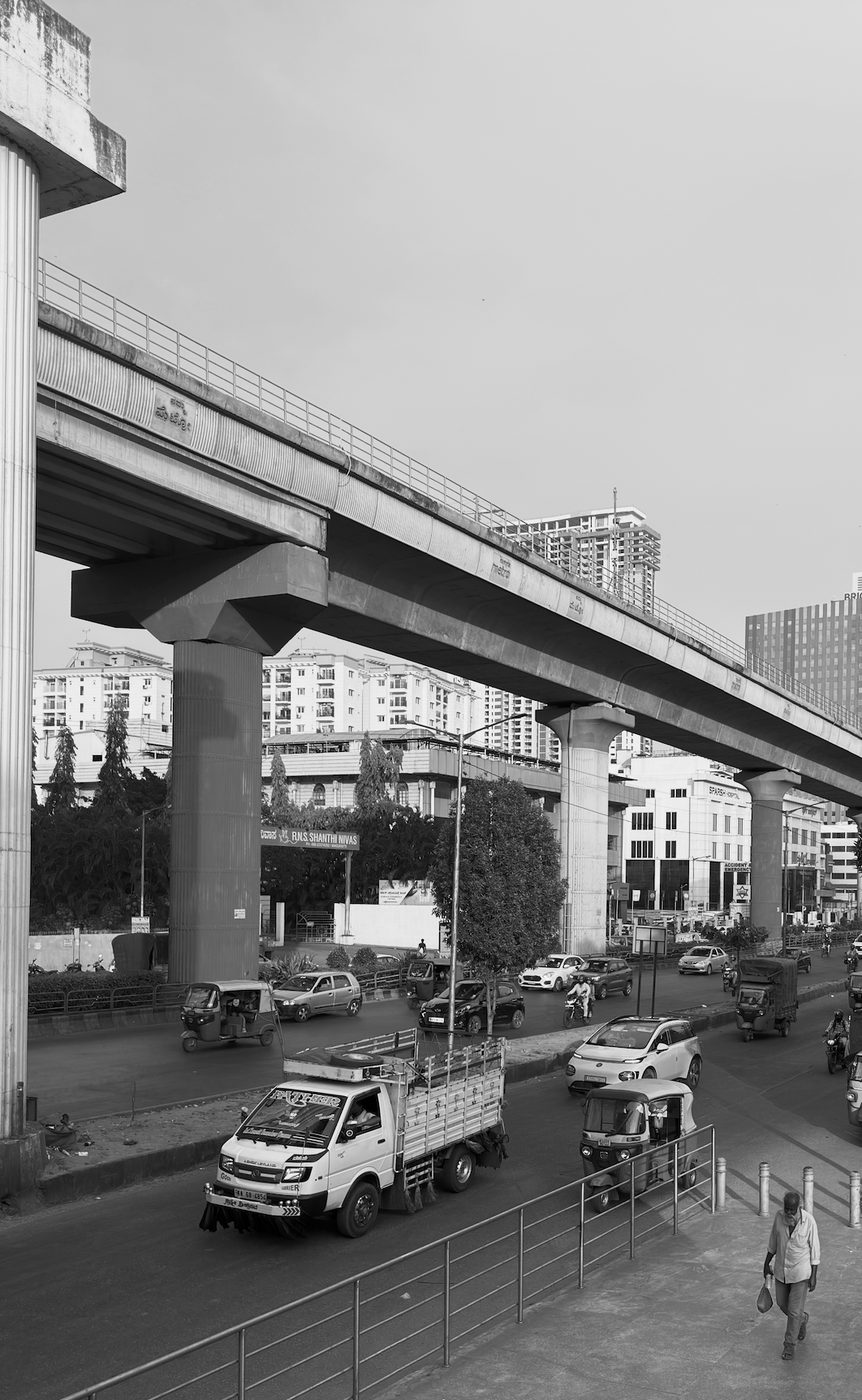 The elevated Bengaluru metro line above a busy street with trucks and auto-rickshaws, in black and white