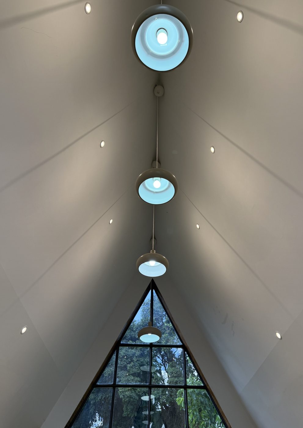 Looking up an A-frame cabin ceiling with pendant lamps leading to a triangular window of trees